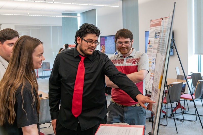 Student in a tie pointing at a research poster while others stand around him