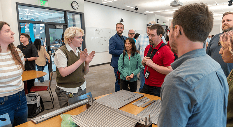 Students presenting to a group of showcase attendees in front of a large table with sheets of metal on it.