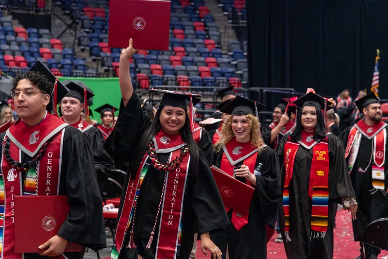 Students walking down the floor of the Toyota Center in regalia. One student is holding their diploma cover above their head.