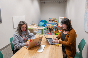 Cristal Sternfeld and Evelyn Martinez-Ostrom working on laptops while their children play with toys in the background.