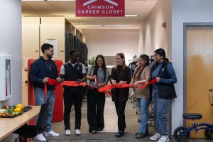 A woman holding large scissors cuts a red ribbon held by five other people under a sign that reads, Crimson Career Closet.