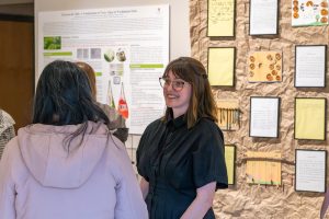 Student Destiny Kuespert speaking with an attendee of the art exhibit opening in front of a large wall of framed student projects.