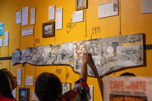 Two people point and look at a photo on a larger artistic timeline on the wall.