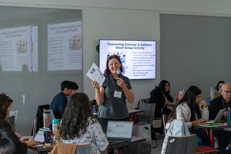 WSU Tri-Cities faculty member holding a piece of paper and microphone around students seated at tables.