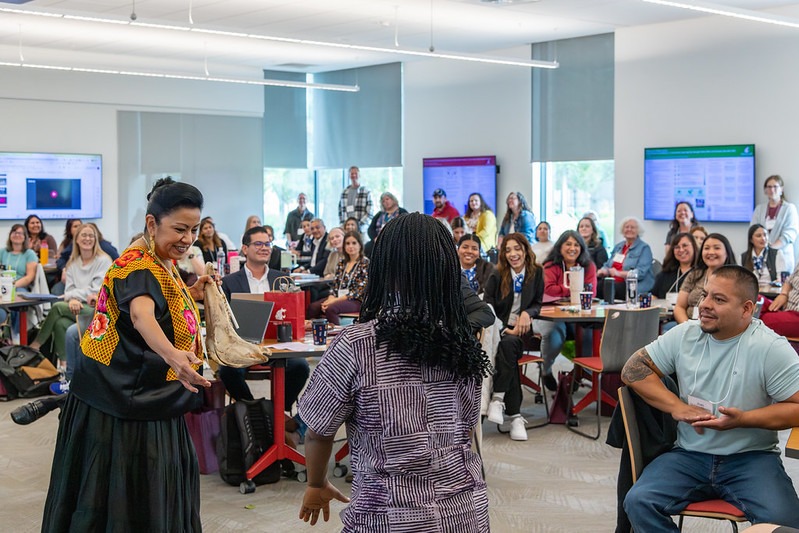 Woman holding a musical instrument inviting another woman to dance with her in front of a room of people.
