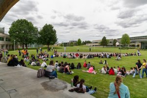 A large group of elementary school students and staff gather around an outdoor basketball court watching three people fly drones.