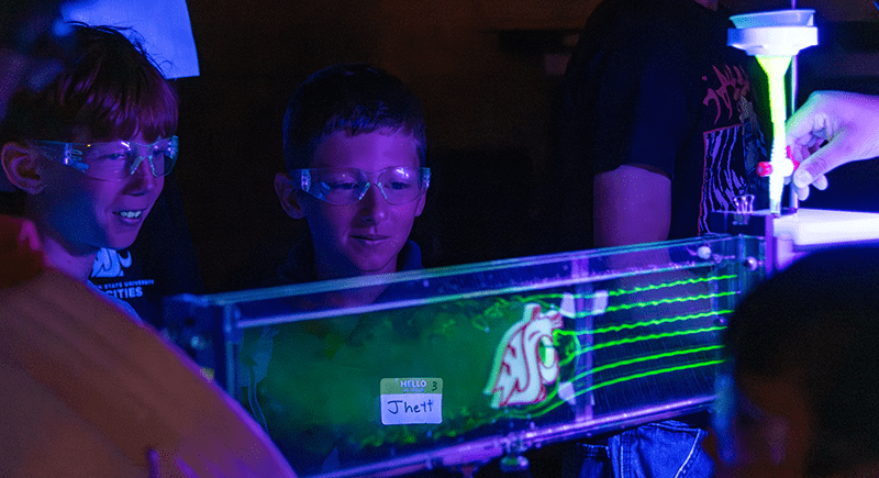 Two children in a dark room lit by UV lights watch as water dyed neon yellow collides with a 3-D printed Coug head in a tank.