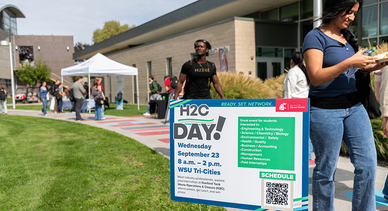 A yard sign stuck in the ground in front of a campus building with a group of students gathered in the background. The yard sign reads, "H2C Day Wednesday September 23 8 a.m. - 2 p.m. WSU Tri-Cities. Meet inudstry professionals, explore jobs/internships at Hanford Tank Waste Operations & Closure (H2C), attend panels, get lunch, and win prizes." The yard sign also features a list of career fields and a QR code to a schedule.
