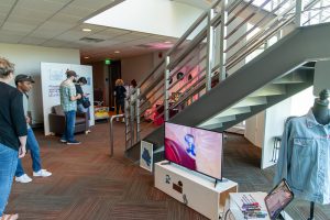 Several people walking by tables displaying artworks including videos on monitors, a denim jacket, and vinyl records.