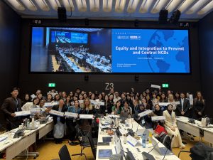 A group of international students standing under a screen that reads, Equity and Integration to Prevent and Control NCDs.