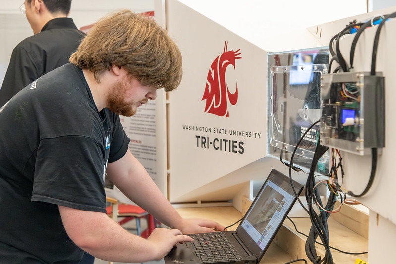 Student working at a laptop in front of a wind tunnel with the WSU Tri-Cities logo painted on it.