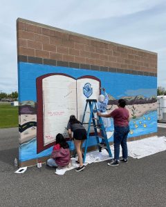 four students paint a mural of a landscape and a book on a brick wall