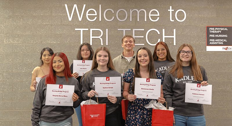 Eight Tri-Tech students holding WSU Tri-Cities Nursing Bridge certificates in front of wall with the letters "Welcome to Tri-Tech" written on it.