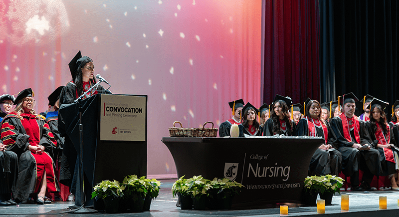 Edith Nateras speaking at a podium on stage with other students and faculty seated behind her.