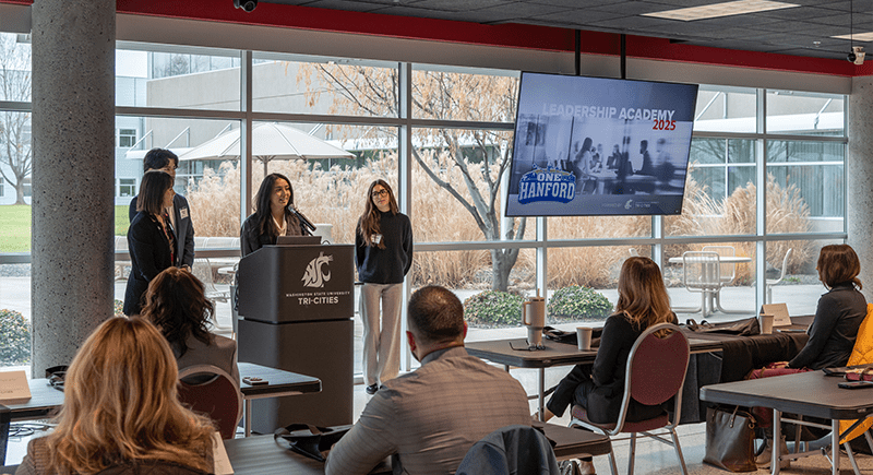 WSU Tri-Cities business student speaks to a group of seated Leadership Academy participants. To her left is a large monitor with the words "Leadership Academy 2025 One Hanford" on it.