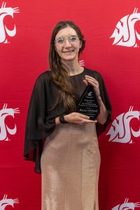 Person standing in front of a crimson Coug head backdrop holding an award that reads, "WSU Tri-Cities Chancellor's Award for Distinguished Career in Leadership presented to Rachel Holland."
