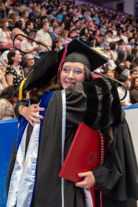 Smiling student wearing graduation regalia and holding a crimson WSU diploma hugs a faculty member.