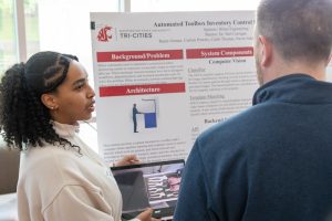 Reem Osman presenting her toolbox project on a laptop to a community member. Behind her is a research poster.