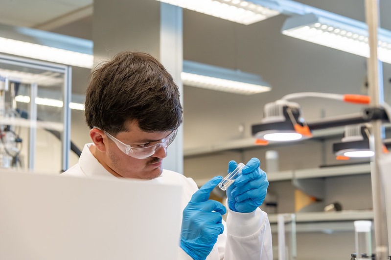 Student wearing a lab coat, gloves, and goggles inspecting a tube of liquid.