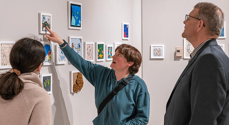 Three people view a wall of small framed artworks in a gallery, with one person pointing to a piece near the top.