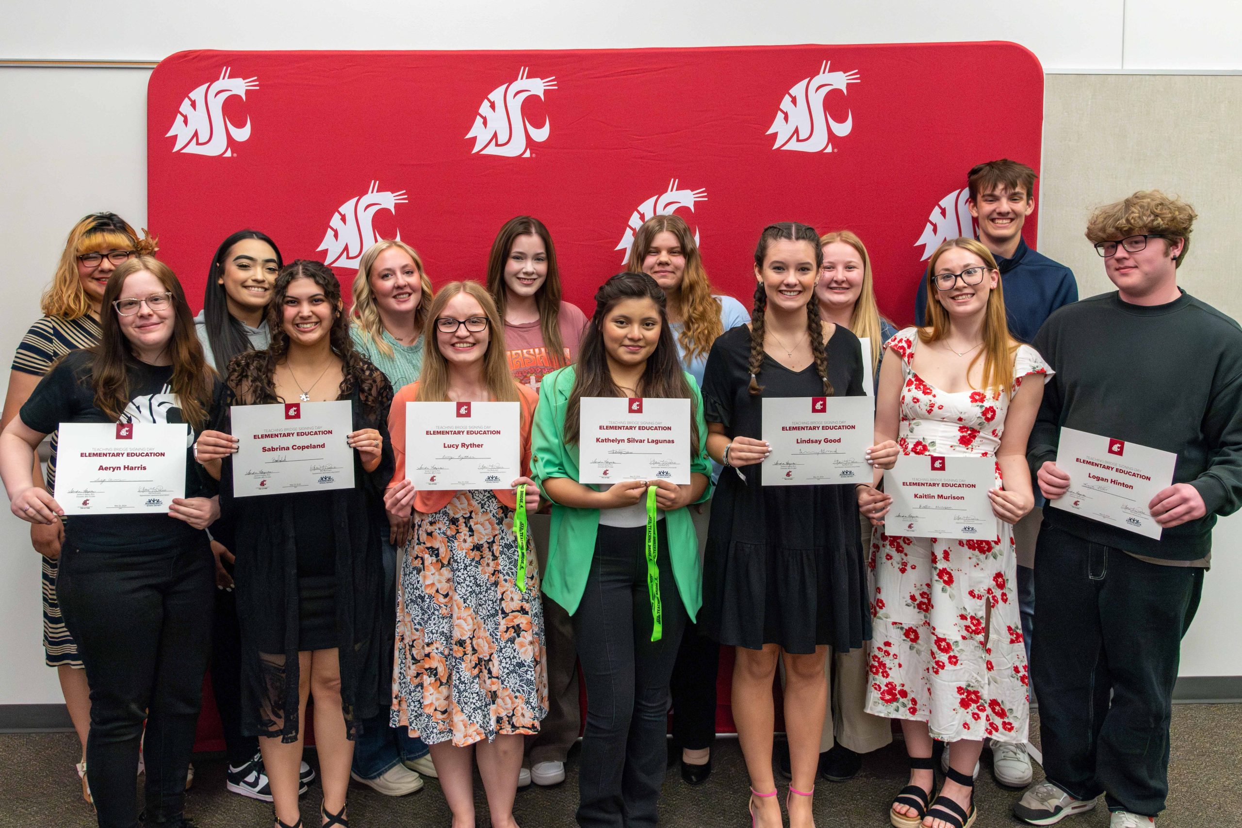 14 Kennewick school district students posing with certificates for a group photo in front of a crimson backdrop with white Coug heads