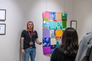 Tyler Hansen speaking to an audience in front of a large electronic textile quilt hanging on the wall.