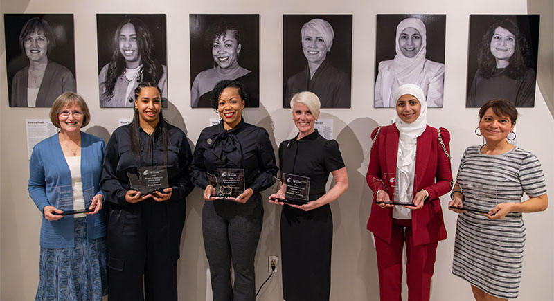 Six Women of Distinction winners holding their awards and standing in front of black and white portraits of themselves