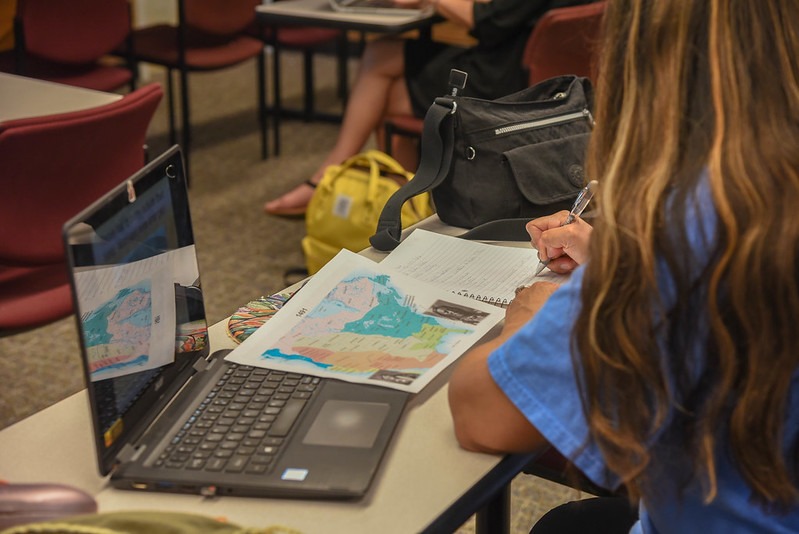 Student at a desk writing in a notebook with a laptop and map.