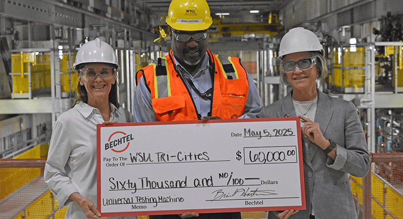 WSU Tri-Cities Chancellor Sandra Haynes, Bechtel Project Director Brian Hartman, and WSU President Elizabeth Cantwell wearing hard hats and protective eye wear in the Hanford site holding a large check that reads “May 5, 2025 Bechtel pay to the order of WSU Tri-Cities $60,000 Universal Testing Machine.”