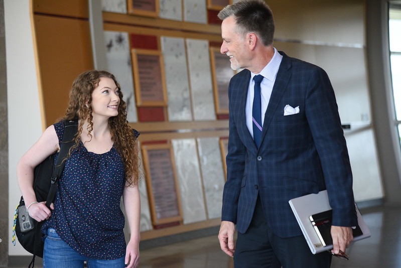 Student with a backpack and professor in a suit speaking in a hallway while walking.