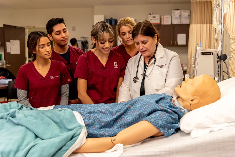 Four students in crimson scrubs and a nursing instructor stand over a mannequin in a hospital bed.