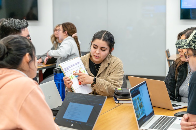 Students seated at a table in a classroom. One student is presenting a figure in a notebook to the others.