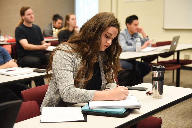 Student seated at a desk in a classroom writing in a notebook.