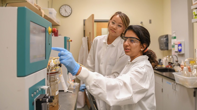 Two students in lab coats and protective eyewear looking at the screen of a machine in a lab.