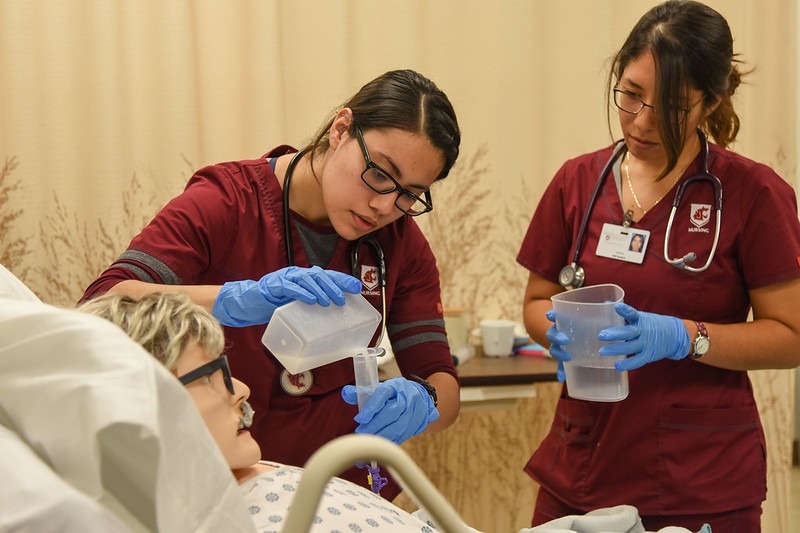 Two nursing students wearing scrubs pouring medicine into a glass by a hospital bed.