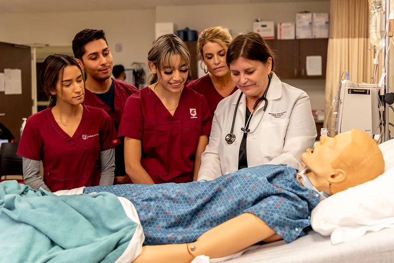 Four WSU Tri-Cities students and instructor dressed in scrubs looking at mannequin on a bed.