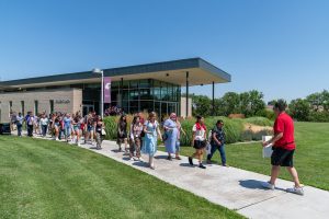 New students walking along a path outside being lead by a man in a crimson shirt holding a large sign.