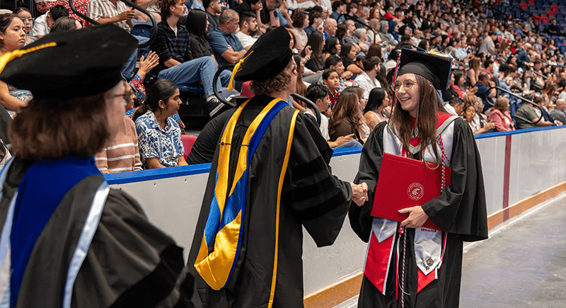 Smiling student wearing graduation regalia and holding a crimson WSU diploma cover shakes a faculty member's hand.