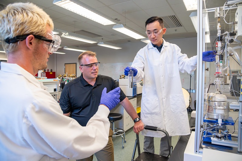 Graduate student handing another student a beaker of liquid while staff member supervises.