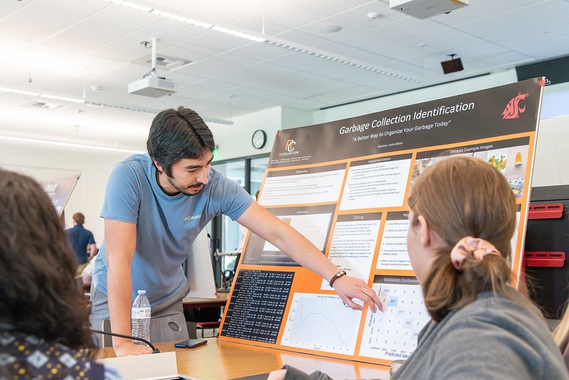Student pointing at a research poster while two other students seated at a table watch.