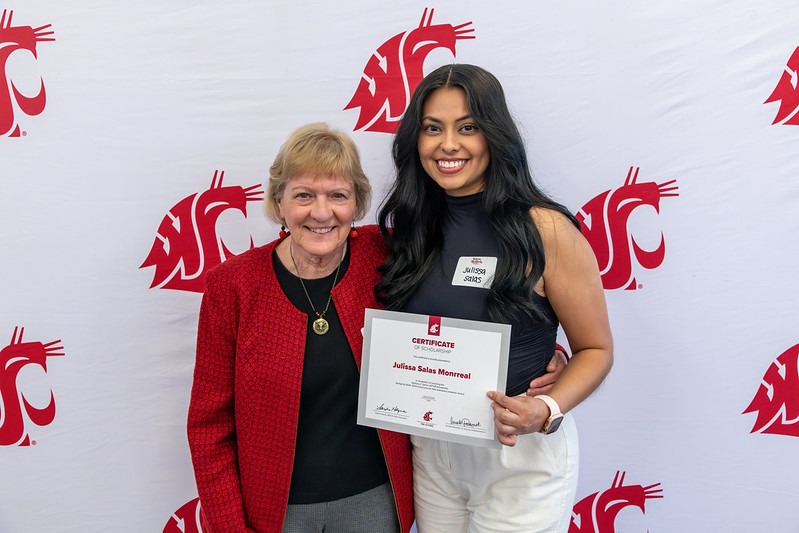 WSU Tri-Cities scholarship recipient and donor standing in front of a crimson and white Coug backdrop. The student is holding a scholarship certificate.
