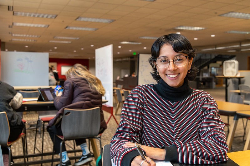 Student smiling at the camera while sitting in the Learning Commons. She is holding a pen and behind her are other students at tables and white boards.