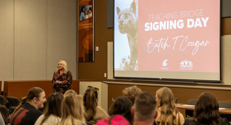 Kennewick School District members seated looking at a projected slide with a photo of Butch and the words "Teaching Bridge Signing Day" along with the WSU Tri-Cities and Kennewick School District logos.