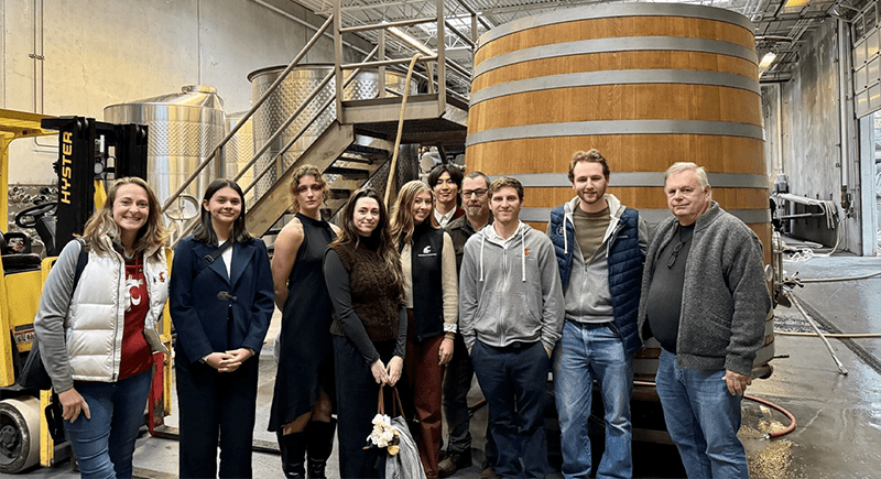 A group of students and faculty posing for a group photo in a wine production facility.