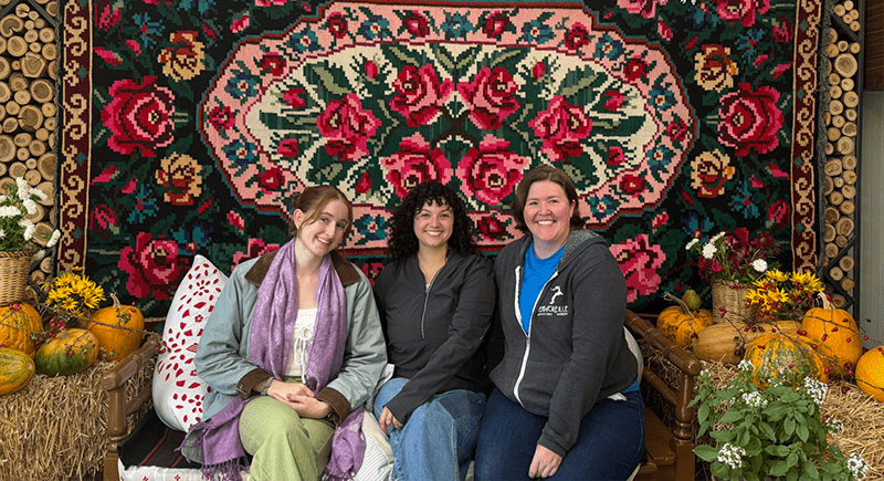 Three students sit in front of a multicolored rug tapestry.