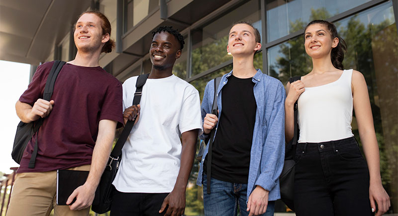 Four college students with backpacks standing in a line and smiling while looking up and to their right.
